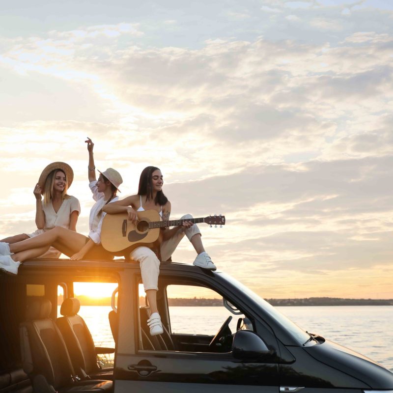 Happy friends with guitar sitting on car roof outdoors at sunset. Summer trip